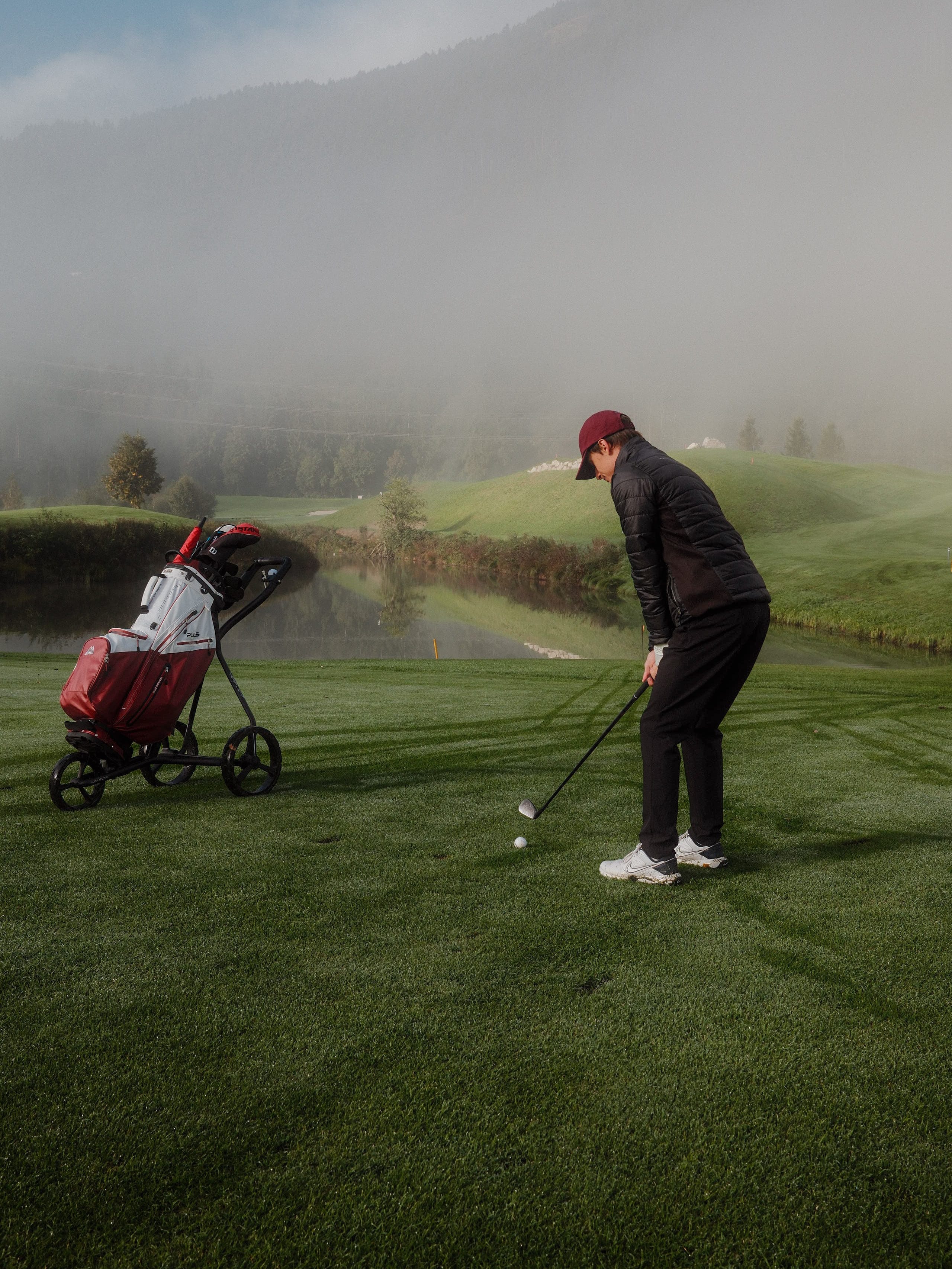 Ein Golfspieler setzt auf einem nebligen Golfplatz zum Schlag an, daneben steht eine Golftasche auf einem Trolley.