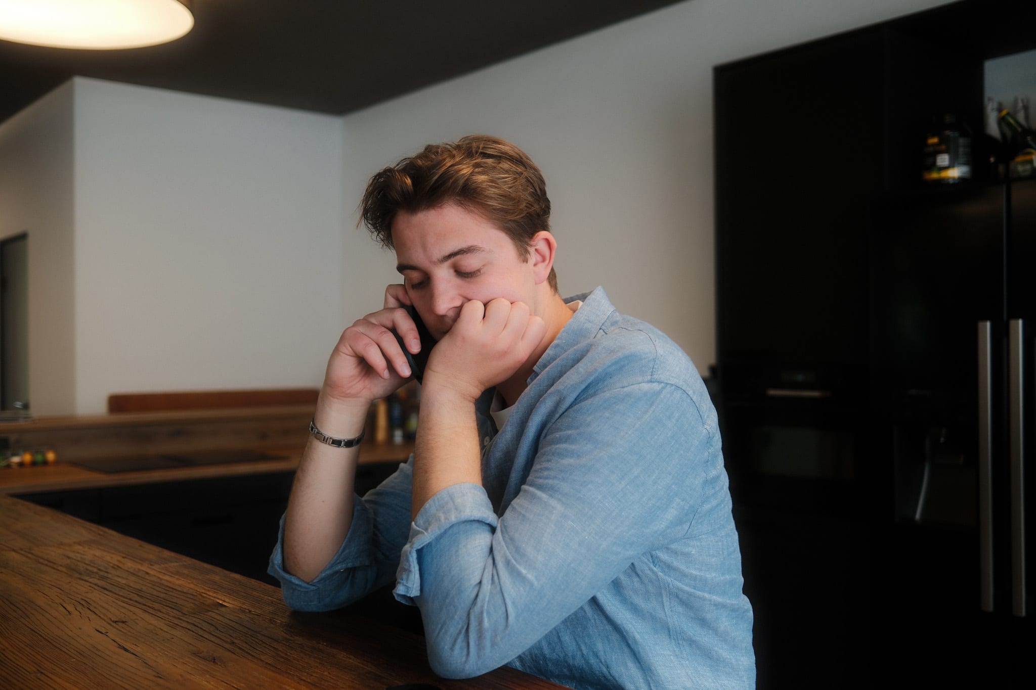 Ein junger Mann sitzt an einer Holztheke, stützt sein Kinn auf eine Hand und hält ein Smartphone ans Ohr.