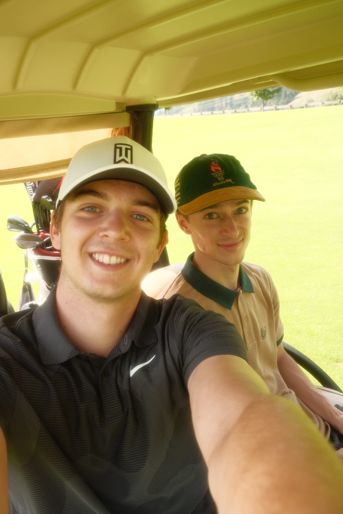 Zwei junge Männer mit Kappen machen ein Selfie in einem Golfwagen auf einem grünen Golfplatz.