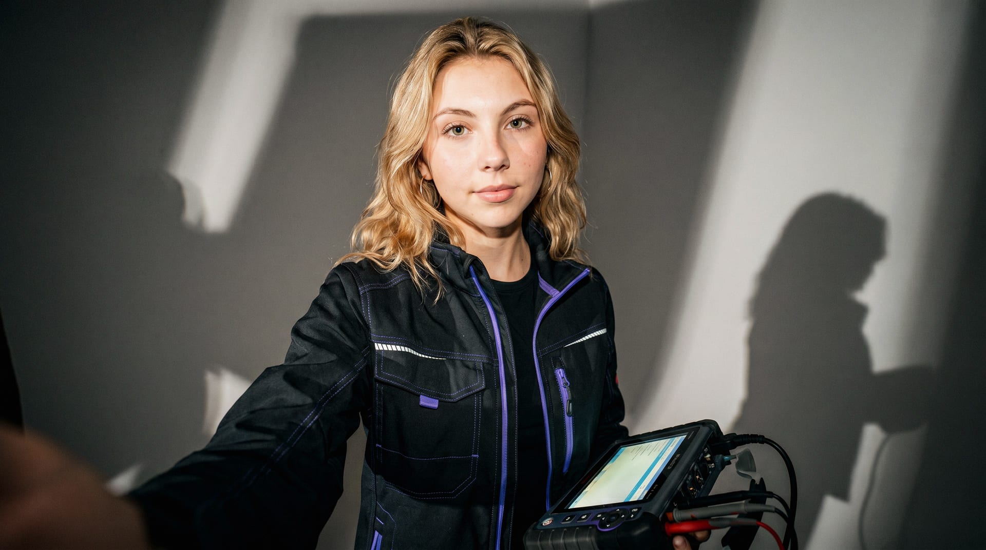 Eine junge Frau in einer Arbeitsjacke mit lila Details hält ein elektronisches Prüfgerät.