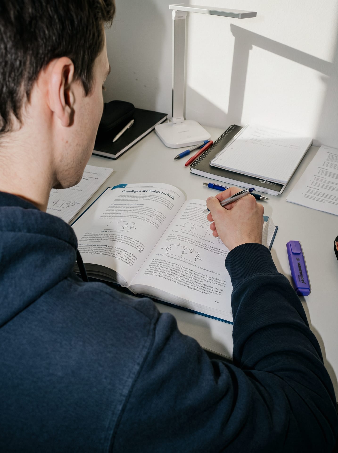Ein Lehrling macht sich am Schreibtisch Notizen in ein Lehrbuch über Grundlagen der Elektrotechnik.